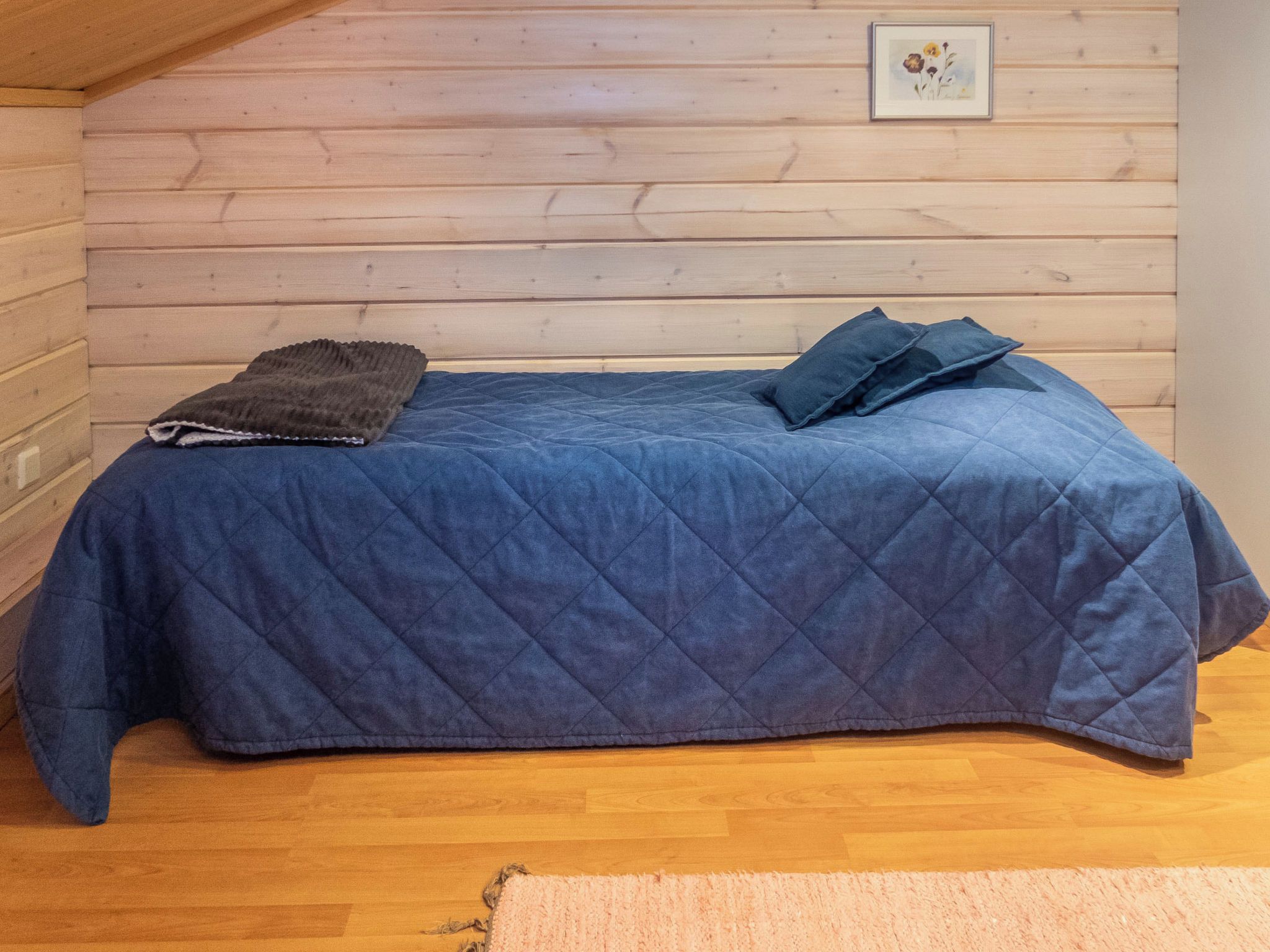 A single bed with a blue quilted bedspread and two pillows, a dark throw blanket, and a wooden floor.