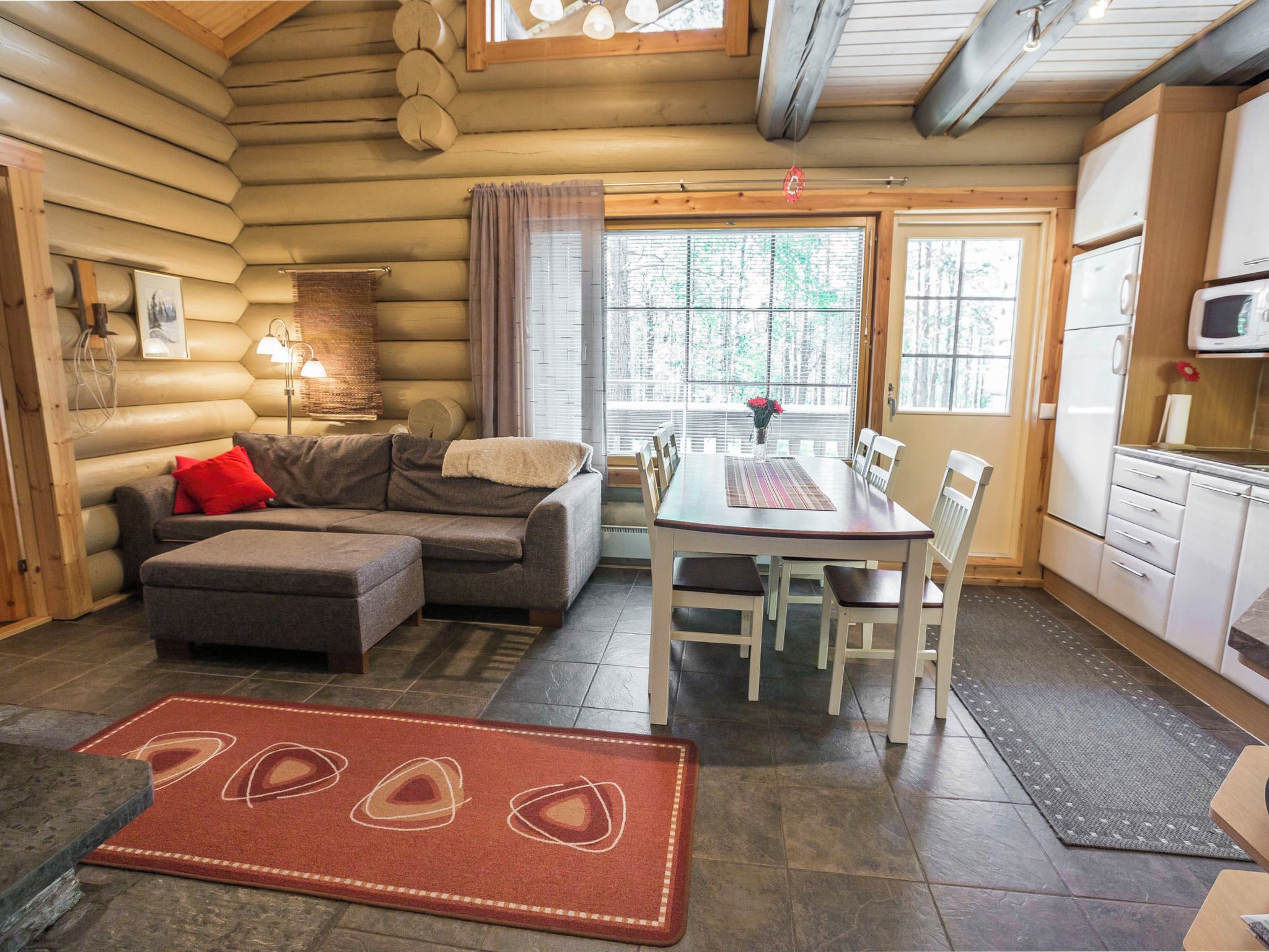 A log cabin interior with a sofa, ottoman, dining table, chairs, kitchen area, and a large window.