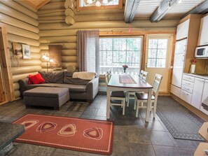 A log cabin interior with a sofa, ottoman, dining table, chairs, kitchen area, and a large window. - Ylläsmaisema 1 by Interhome (Ylläsjärvi)