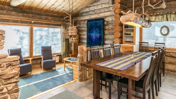 A rustic log cabin interior with a dining area, stone fireplace, and large windows.