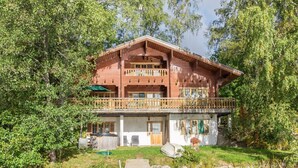 Een houten chalet met twee verdiepingen en een balkon, omgeven door weelderige begroeiing en een steiger naar een vijver.