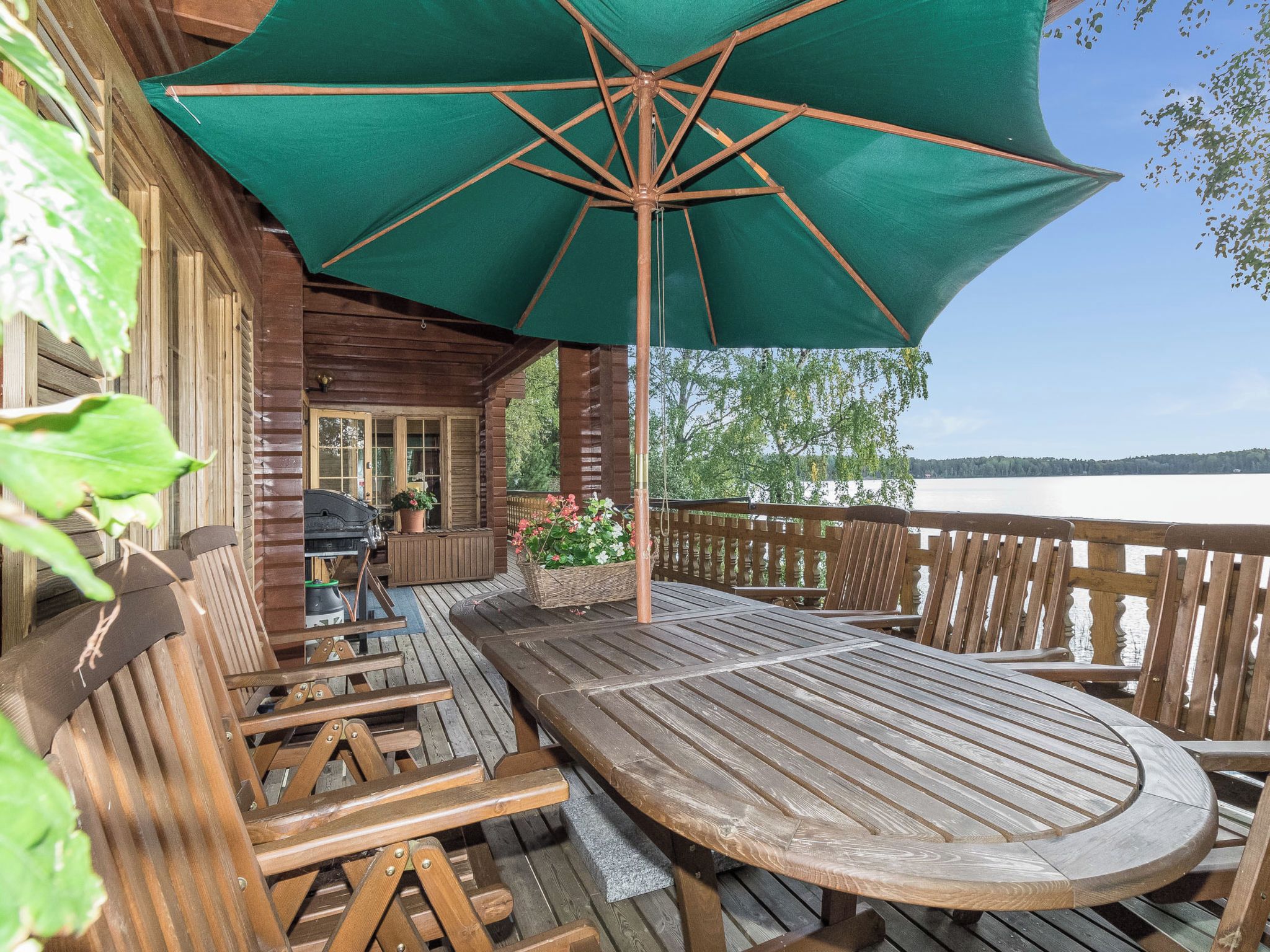 Eine Holzterrasse mit Tisch und Stühlen, einem grünen Sonnenschirm und Blick auf einen See.