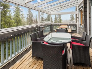 Et overdækket udendørs opholdsområde med fletmøbler og et glasbord, med udsigt over et skovområde.