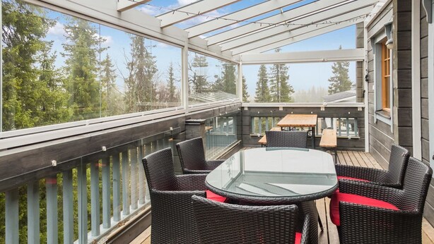 A covered outdoor patio with wicker furniture and a glass table, overlooking a forested area.