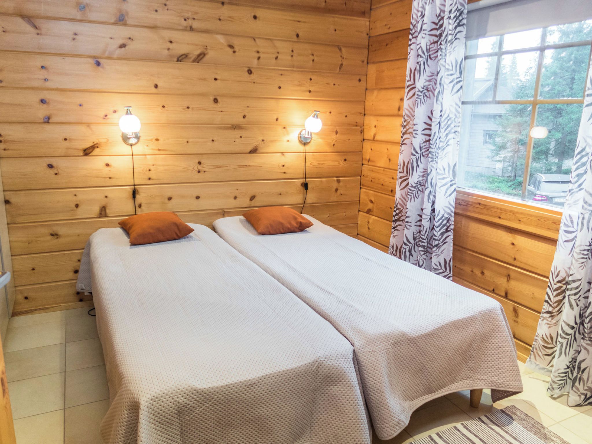 A room with wooden walls, two beds with brown bedding, a window with curtains, and two wall-mounted light fixtures.
