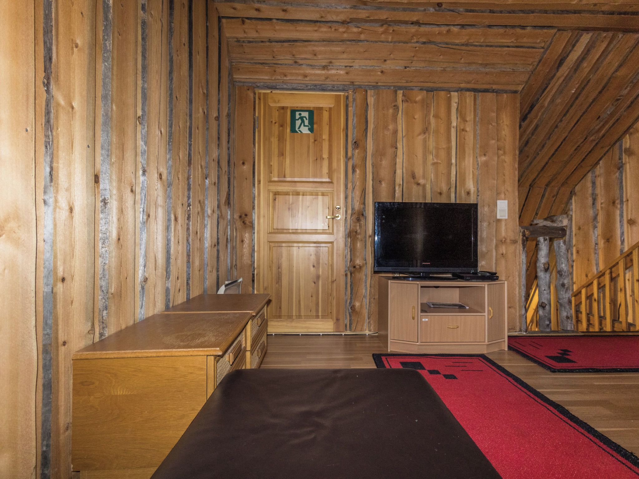 A wooden interior with a bench, a television on a cabinet, and a door with an emergency exit sign.