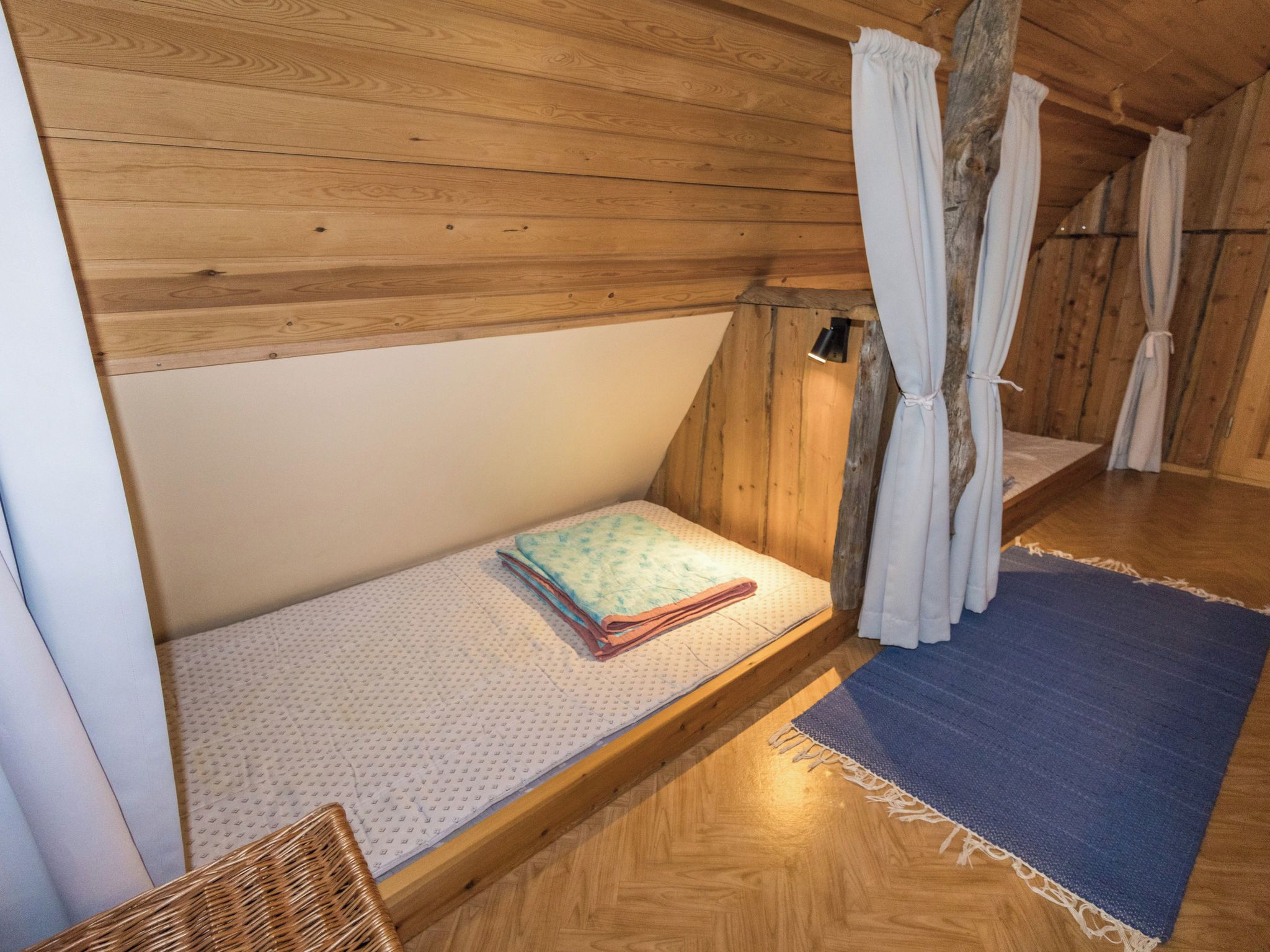 A small, wooden, attic-style room with a single bed, a chair, and a window with curtains.