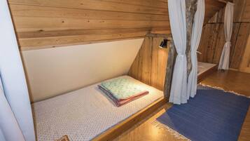A small, wooden, attic-style room with a single bed, a chair, and a window with curtains.
