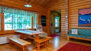 A log cabin interior with a wooden dining table, benches, a sofa, and a large window with a view of trees.