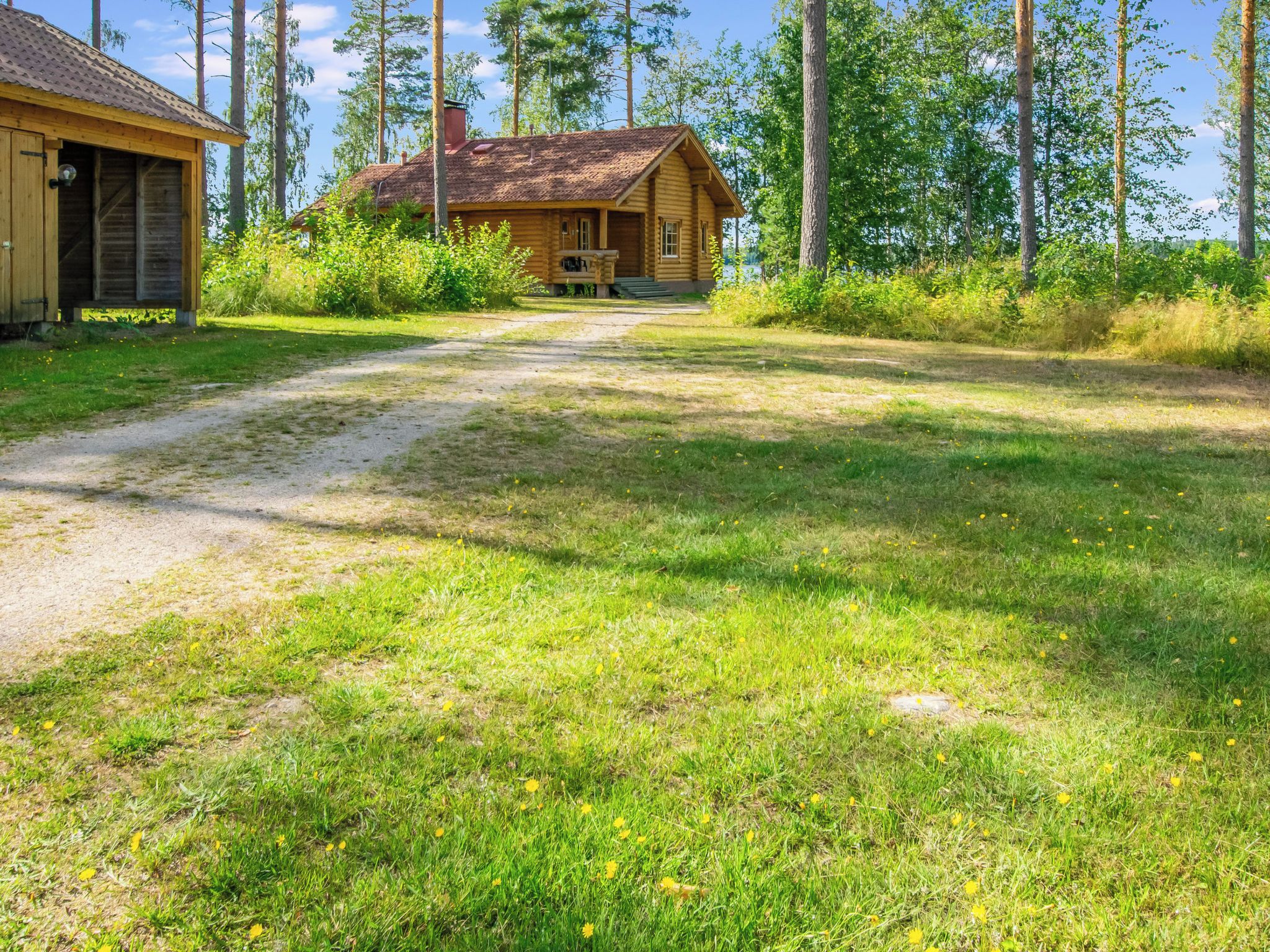 Une cabane en bois, accessible par un chemin de terre, entourée d’arbres et d’herbe.