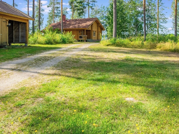 Eine Holzhütte mit einem Feldweg, der davor hindurchführt, umgeben von Bäumen und Gras.
