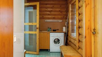 Interior de una cabaña pequeña y de madera, con lavadora, una mesita y un banco.