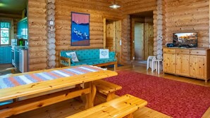 A log cabin interior with a wooden dining table, a sofa, a red rug, a painting, and a television on a cabinet.