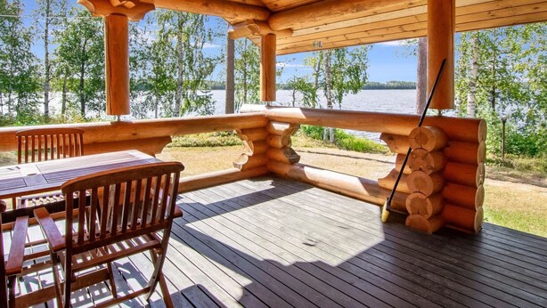 A wooden deck with a table and chairs, overlooking a lake with trees and a clear sky.