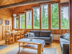 Ein gemütliches Blockhaus-Interieur mit Essbereich, einer Couch und großen Fenstern mit Blick auf Bäume.