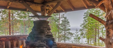 Ein Steinkamin mit Feuer, umgeben von Holzbalken und Blick auf Wald und Wasser.
