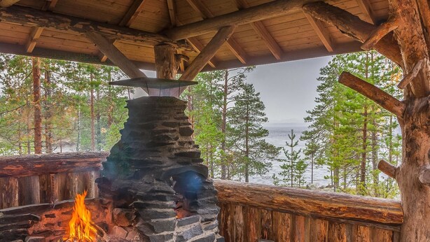 Una chimenea de piedra con fuego, rodeada de leña y una estructura de madera rústica con vista a un lago y árboles.