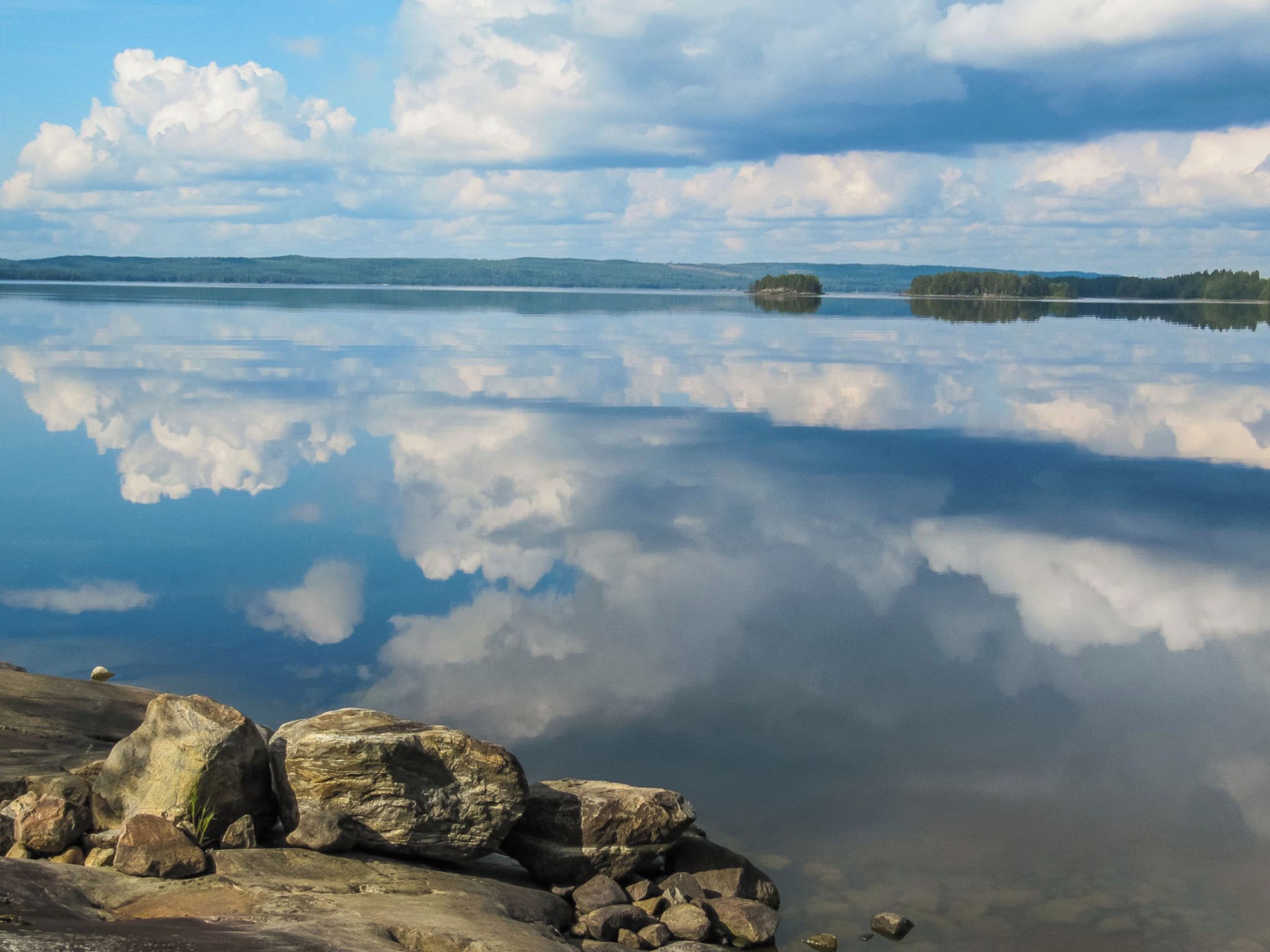 Un lago tranquilo con reflejos nítidos del cielo y las nubes, rodeado de rocas y árboles distantes.