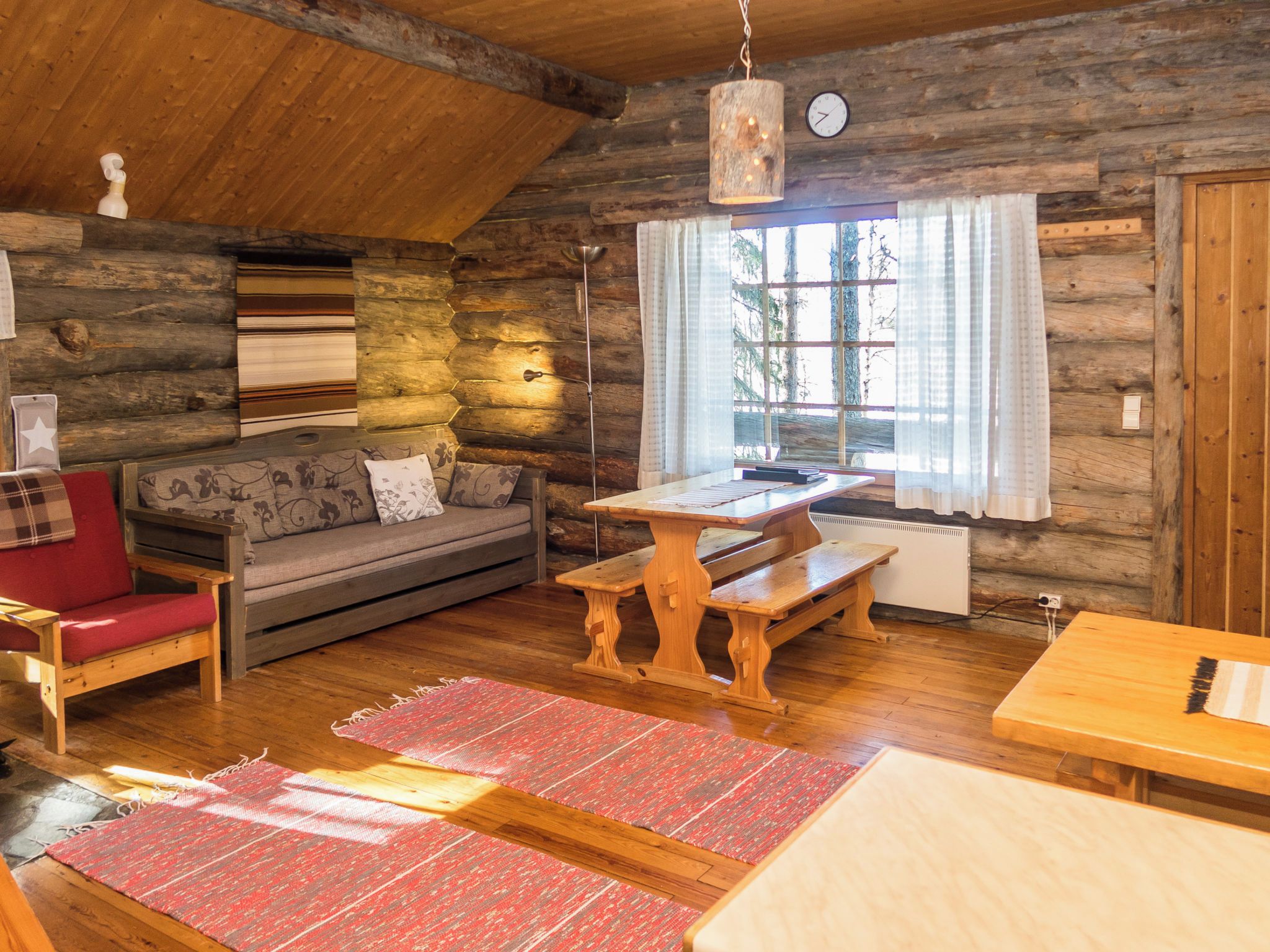 A rustic cabin interior with a wooden ceiling, a sofa with patterned cushions, a dining table with benches, and a large window with curtains.