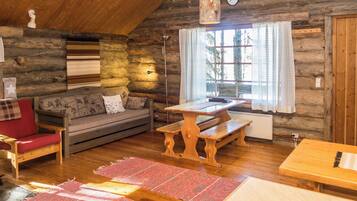 A rustic cabin interior with a wooden ceiling, a sofa with patterned cushions, a dining table with benches, and a large window with curtains.