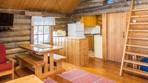 Interior d'una cabana rústica amb taula de dinar de fusta, una cadira vermella, televisió penjada a la paret, zona de cuina amb armaris de fusta i una escala de fusta.