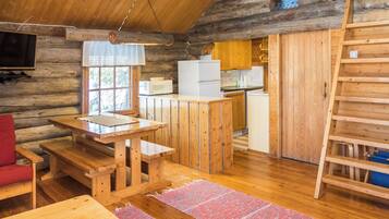 A log cabin interior with a wooden dining table, a red chair, a kitchen area with a refrigerator and microwave, and a wooden ladder.