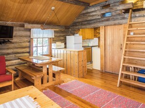 Interior de una cabaña de troncos con una mesa de comedor de madera, una silla roja, una zona de cocina con refrigerador y microondas, y una escalera de madera.