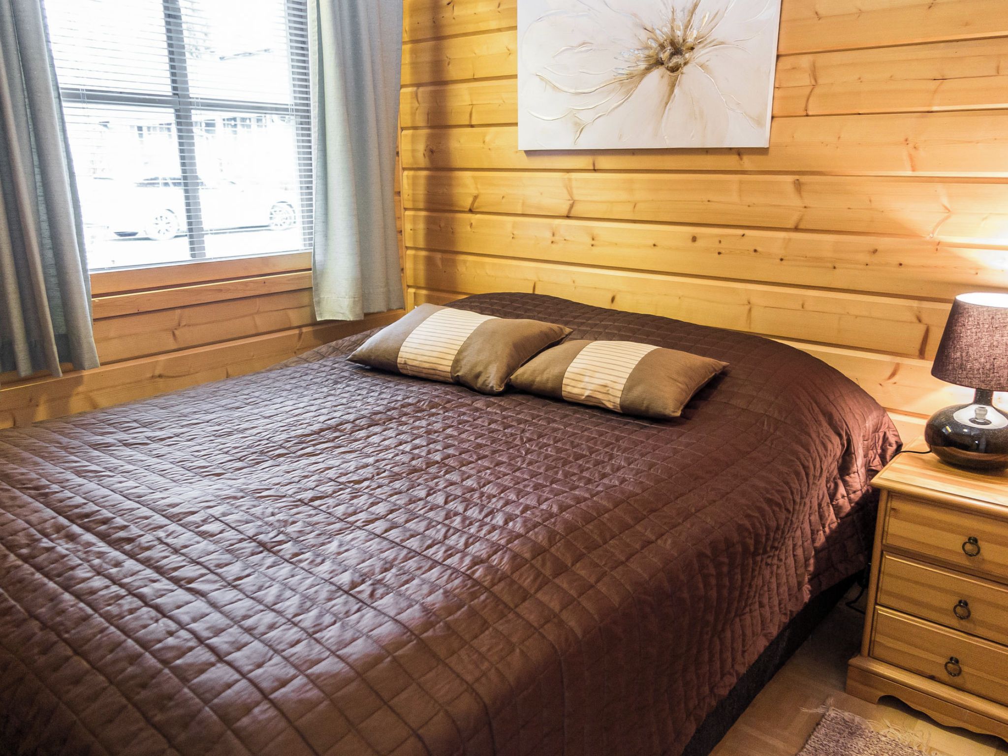 Habitación con paneles de madera, una cama, una mesita de noche, una lámpara y un ventanal con cortinas.