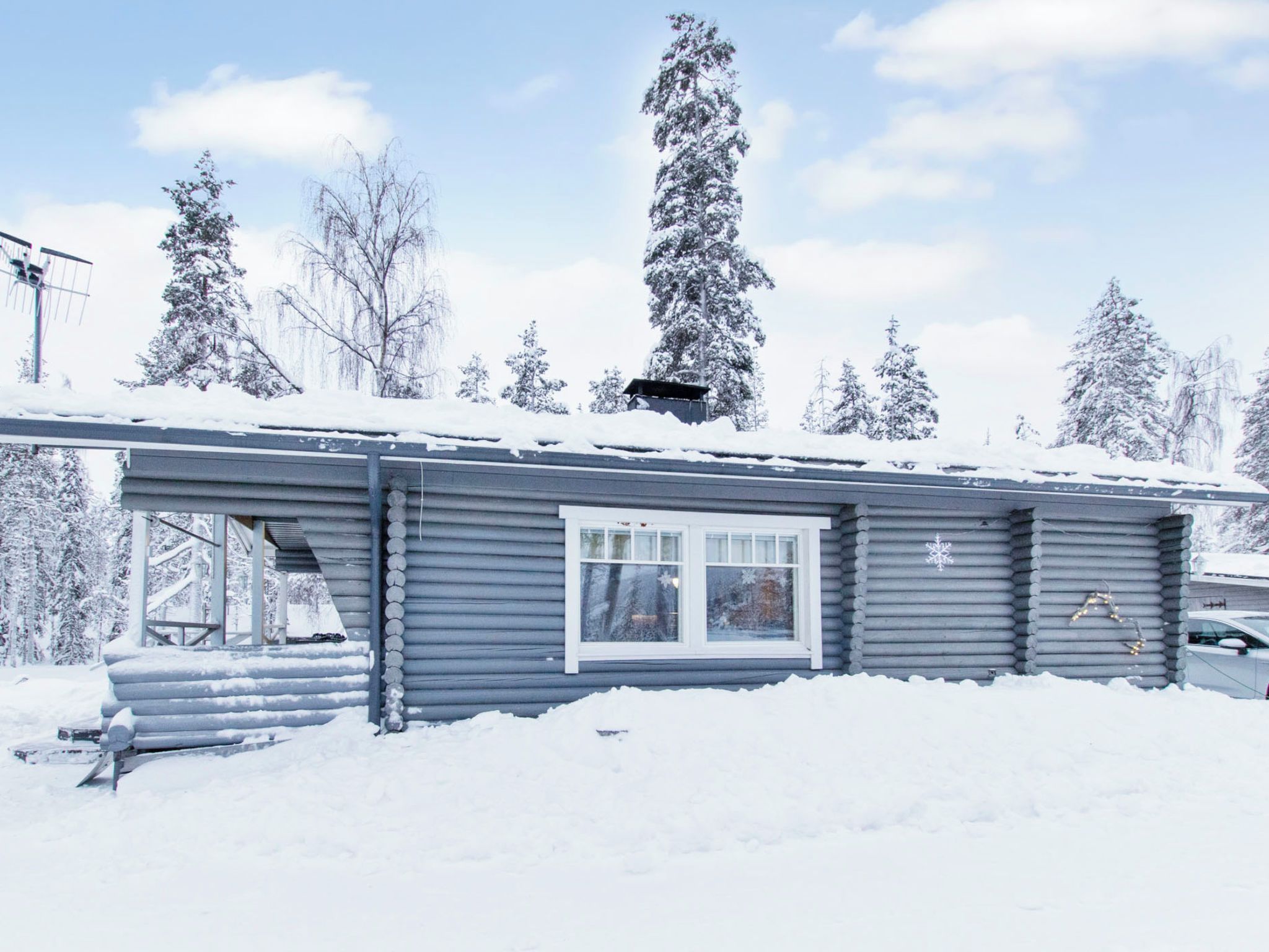 Une cabane en rondins avec une fenêtre et un toit recouvert de neige, entourée de neige et d’arbres.