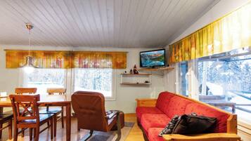 Een gezellige woonkamer met een houten plafond, een rode bank, een houten eettafel met stoelen, een televisie op een plank en grote ramen met gordijnen.