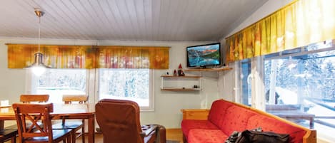 A cozy living room with a wooden ceiling, a red sofa, a wooden dining table with chairs, a television on a shelf, and large windows with curtains.