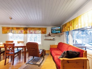 A cozy living room with a wooden ceiling, a red sofa, a wooden dining table with chairs, a television on a shelf, and large windows with curtains. - Jousimaja by Interhome (Kolari)