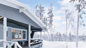 Een met sneeuw bedekte houten blokhut met een veranda en een raam met uitzicht op een besneeuwd bos.