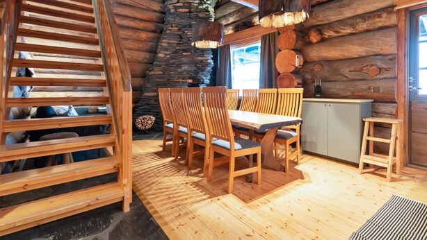 Ein Blockhaushaus-Interieur mit einer Holztreppe, einem Steinkamin, einer Essecke mit Holzsesseln und einer Küchenzeile.