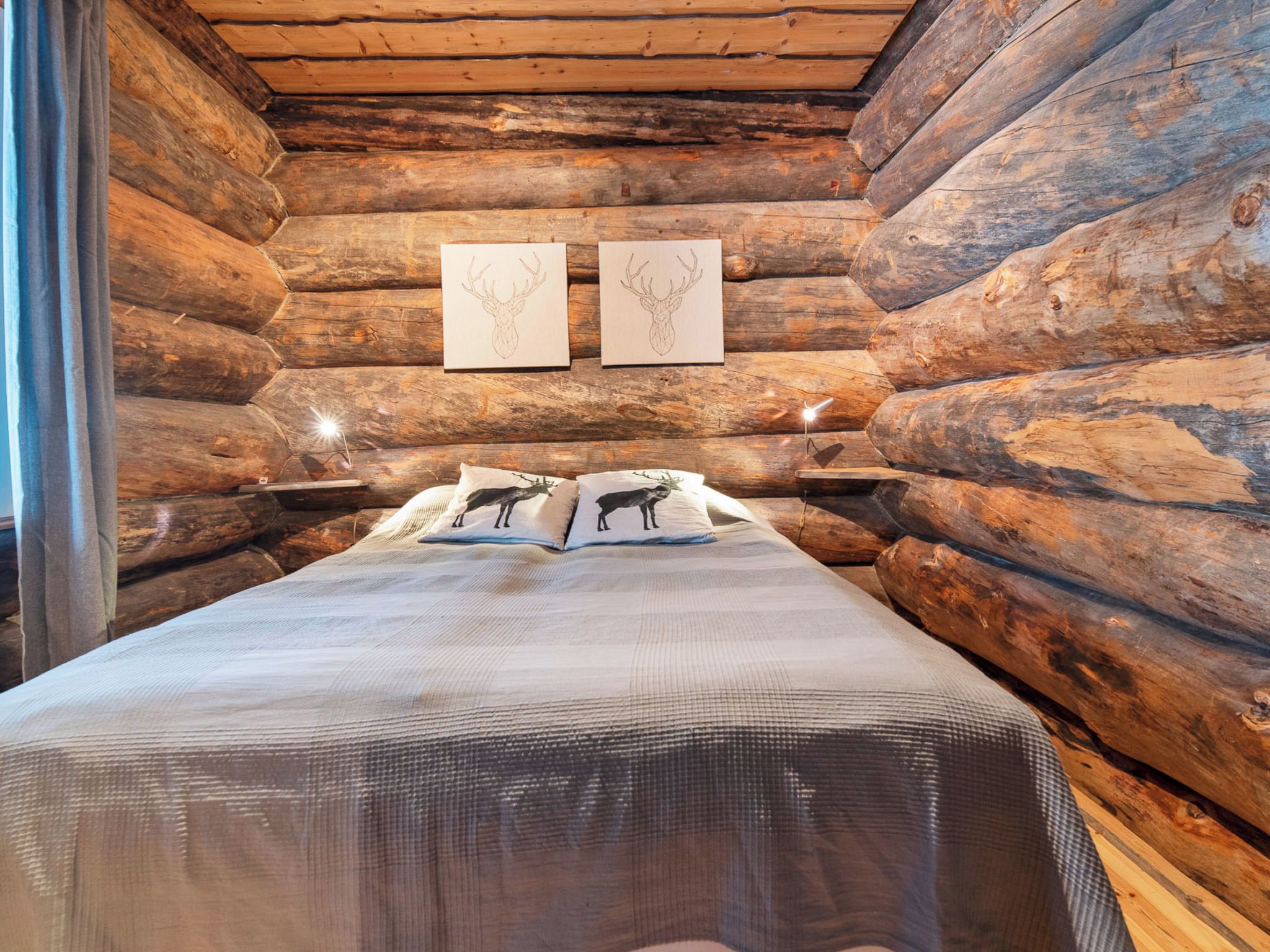 A log cabin bedroom with a bed, two wall-mounted lamps, and two framed deer head paintings.