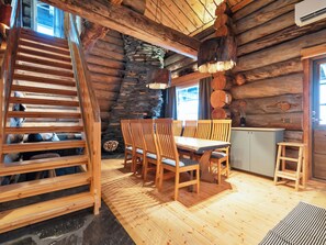 Interior de una cabaña de troncos con una escalera de madera, una zona de comedor con sillas y mesa de madera, una pared de piedra y una cocina.