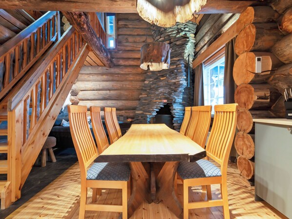 Interior de una cabaña de troncos con una mesa de comedor de madera, sillas, chimenea de piedra y una escalera.