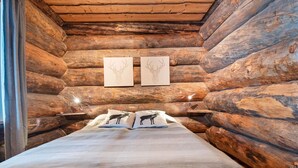 Ein Schlafzimmer in einer Blockhütte mit einem Bett, zwei an der Wand befestigten Zeichnungen und einem Nachttisch mit Lampe.