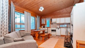A cozy cabin-style room with a wooden ceiling, a sofa, a dining table, and a kitchen area with white cabinets and appliances.