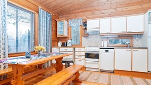 A wooden cabin interior with a dining table, benches, a kitchen with white cabinets, and a window with blinds.