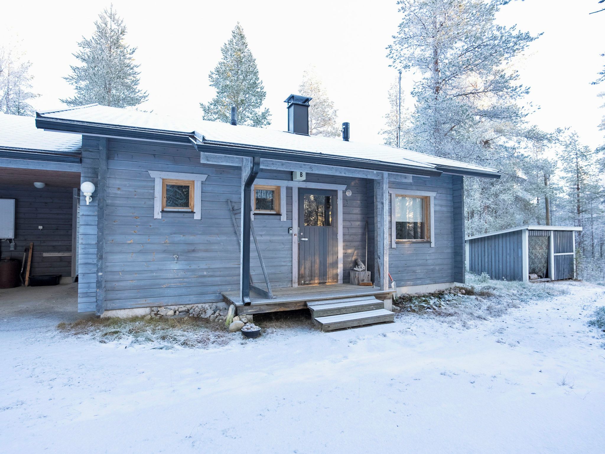Une cabane en bois avec un porche à l’avant, une cheminée et un petit abri de jardin, le tout situé dans un paysage enneigé.