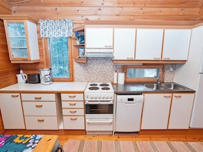A compact kitchen with white cabinets, a stove, a dishwasher, and a window with a view of trees. - Pohjaskoti b by Interhome (Kuusamo)