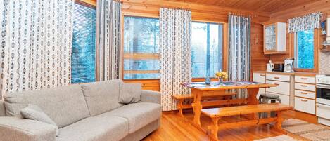 Interior acolhedor de cabana com sofá, mesa de jantar de madeira, área de cozinha e janelas amplas com cortinas.