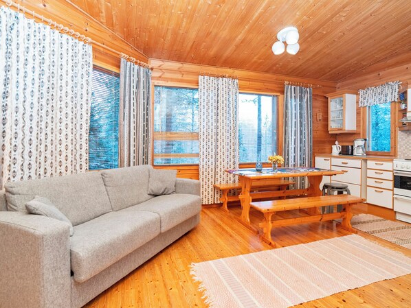 A cozy cabin interior with a sofa, wooden dining table, kitchen area, and large windows with curtains. - Pohjaskoti b by Interhome (Kuusamo)