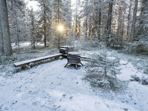 A snowy forest campsite with a metal stove, a wooden bench, and a picnic table. - Pohjaskoti b by Interhome (Kuusamo)