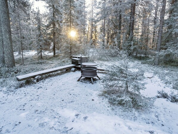 A snowy forest campsite with a metal stove, a wooden bench, and a picnic table. - Pohjaskoti b by Interhome (Kuusamo)