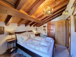 A bedroom with a large bed, wooden beams, a chandelier, a wooden wardrobe, and a nightstand with a lamp. - Haus Rollin by Interhome (Zermatt)