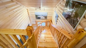 Un escalier en bois à l’intérieur d’une cabane en rondins, avec vue sur une montagne enneigée et sur une autre cabane, visibles à travers une fenêtre.