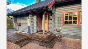Une cabane en rondins avec une terrasse en bois, une porte rouge et une fenêtre dotée d’un cadre jaune.