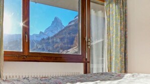A room with a bed, a window with a view of mountains, and a curtain.